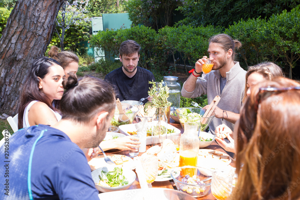 Serious male and female friends having dinner outdoors. Young women and men eating salads and drinking juice in garden. Outdoor party concept Serious male and female friends having dinner outdoors. Young women and men eating salads and drinking juice in garden. Outdoor party concept