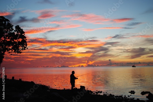 Silhouette of an unrecognizable man casting a fishing pole in the waters with a blood-red sunset in the background.