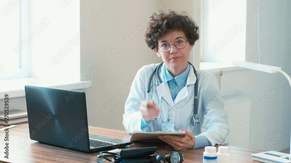 Up-section shot of female doctor filling out medical documents, then raising her eyes to the camera with serious glance Up-section shot of female doctor filling out medical documents, then raising her eyes to the camera with serious glance