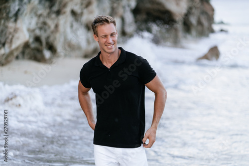 Sexy smiling man in black t-shirt resting on beach, ocean waves at background. 