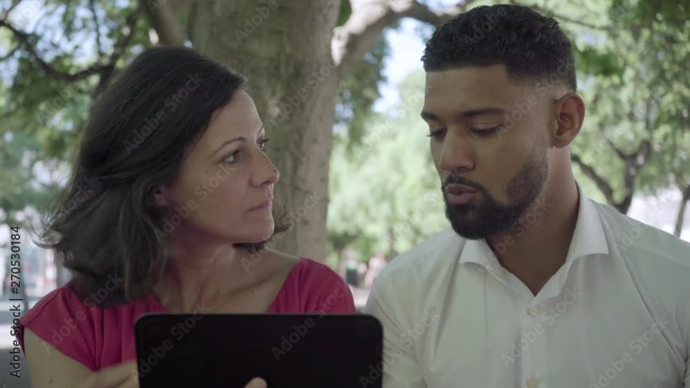 Focused employees looking at tablet and talking outdoor. Concentrated middle aged Caucasian woman and African American man discussing some business issues in summer park. Communication concept