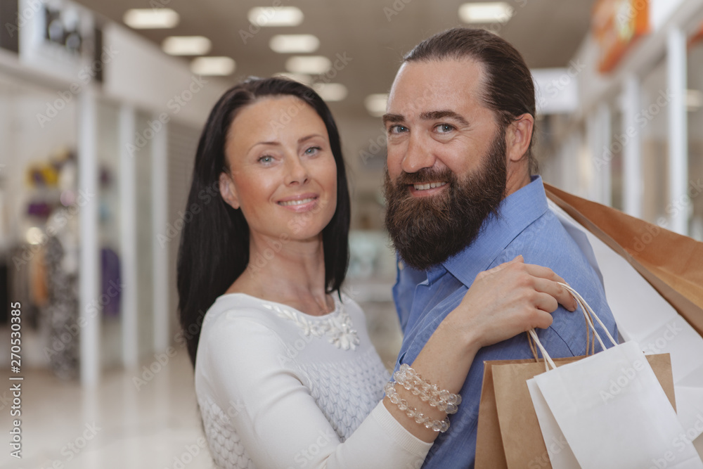Bearded handsome mature man hugging his gorgeous wife, smiling to the camera at the shopping mall. Happy mature couple cuddling after shopping at the mall together, copy space Bearded handsome mature man hugging his gorgeous wife, smiling to the camera at the shopping mall. Happy mature couple cuddling after shopping at the mall together, copy space