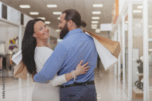 Beautiful happy mature woman smiling to the camera over her shoulder  embracing husband at the shopping mall. Rear view of a couple carrying shoping bags  hugging at the shopping center