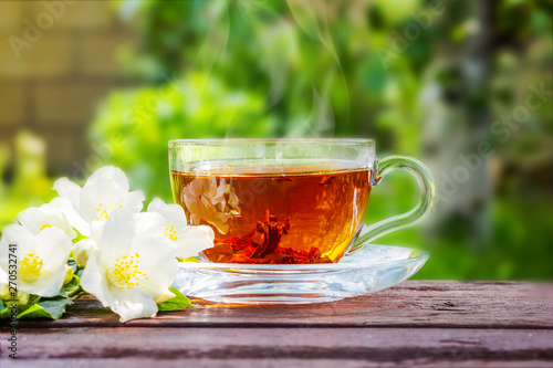 Glass cup with tea and jasmine twig on wooden table, on blurred background