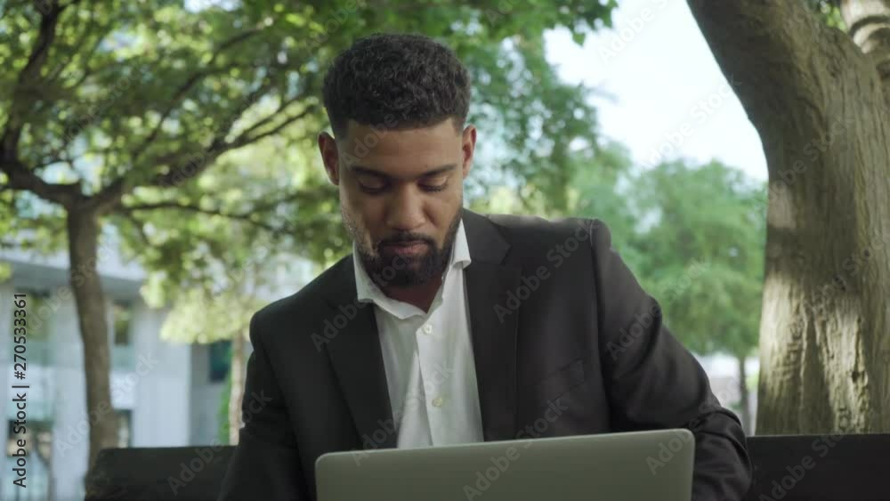 Focused young businessman working with laptop in sunny park. thoughtful African American man wearing formal suit sitting on bench with laptop. Technology concept Focused young businessman working with laptop in sunny park. thoughtful African American man wearing formal suit sitting on bench with laptop. Technology concept