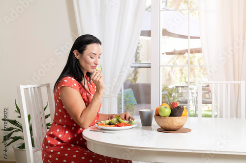 Beautiful brunette woman sitting wearing a red and white polka dot dress at dining table eating a healthy salad and drinking coffee or tea with a bowl of fruits on table with garden in the background