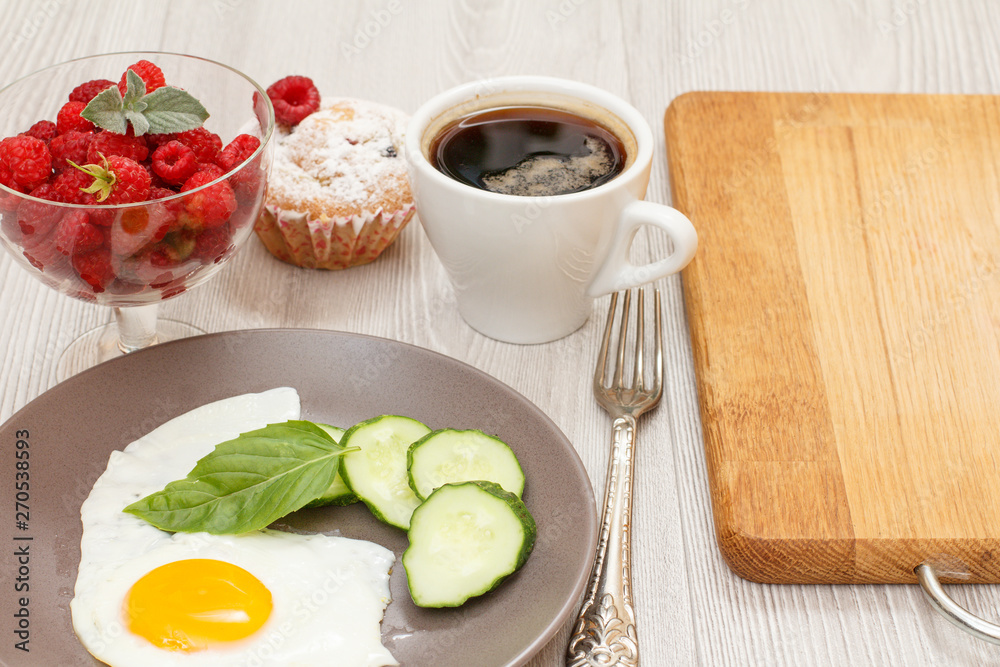 Plate with fried egg, cut cucumber and leaf of basil, glass bowl with raspberries, cup of coffee, fork and muffin. Plate with fried egg, cut cucumber and leaf of basil, glass bowl with raspberries, cup of coffee, fork and muffin.