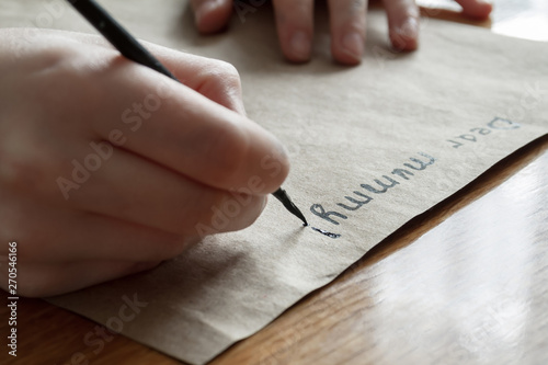 A small girl writes a letter to dear mummy by black paint on brown paper. Closeup, selective focus