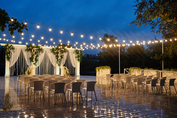 Night wedding ceremony with arch, orchid flowers, chairs and bulb lights in forest outdoors, copy space. Wedding decorations