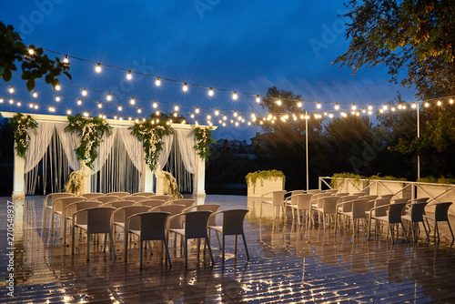 Night wedding ceremony with arch, orchid flowers, chairs and bulb lights in forest outdoors, copy space. Wedding decorations