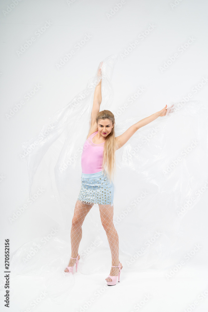 Naklejka premium Beautiful girl with blond hair in a pink t-shirt, blue skirt and mesh white pantyhose dancing with plastic wrap on a white background