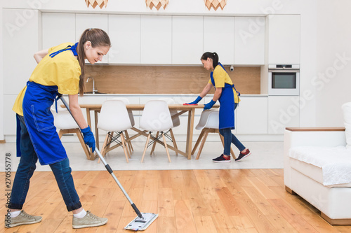 professional cleaning service. Two women in working uniform, in aprons, divide the cleaning of the kitchen of a private house, cottage. washing the refrigerator, tap, sink. Wash floor.