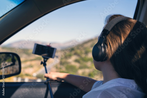 Blogger girl in the headphones taking selfie picture or video using smartphone and selfie stick inside of car.