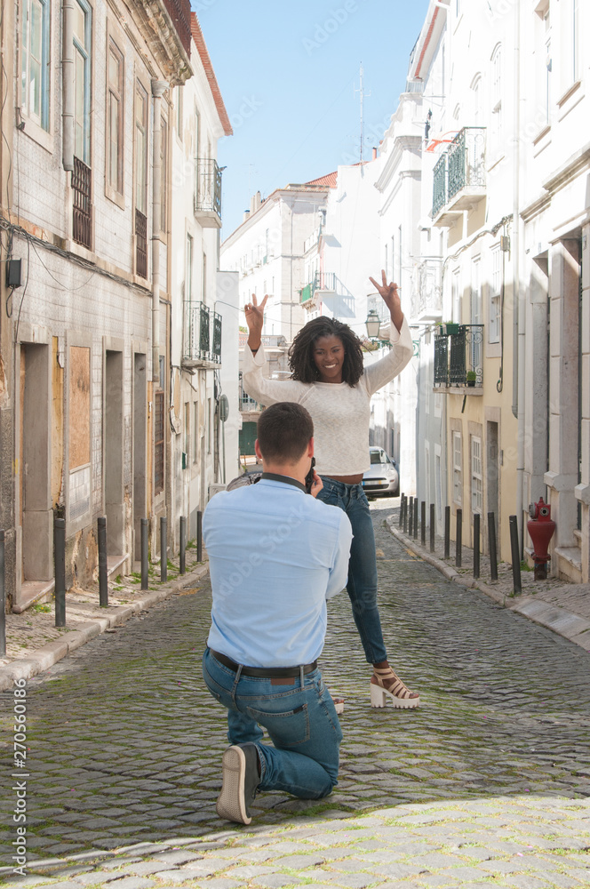 Man taking photo of playful black woman outdoors. Woman showing victory signs and man standing back to camera in city street. Tourism concept. Man taking photo of playful black woman outdoors. Woman showing victory signs and man standing back to camera in city street. Tourism concept.