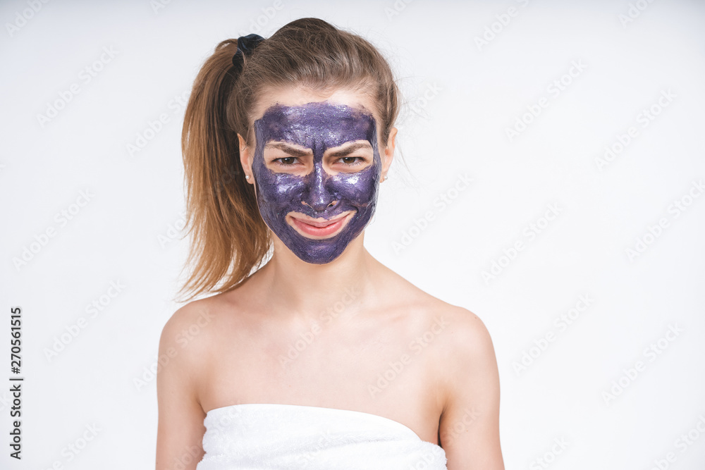 emotional portrait of a young beautiful european girl in violet cosmetic mask on her face. Wrapped in a towel. European, 22 years old, white, tail on the head, blank background, looking straight emotional portrait of a young beautiful european girl in violet cosmetic mask on her face. Wrapped in a towel. European, 22 years old, white, tail on the head, blank background, looking straight