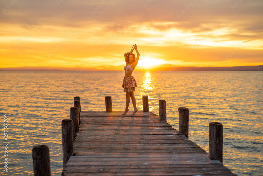 Fototapeta premium Happy woman enjoying the summery sunset at the lake on her vacation. The woman in bra and a short summer skirt laughs and puts her arms in the direction of the sky