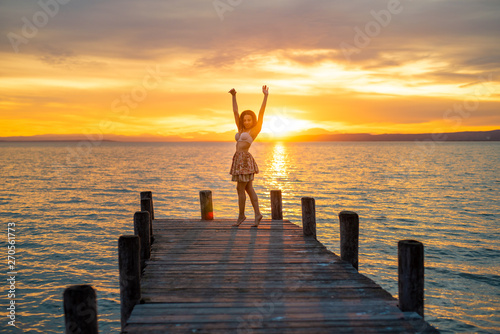 Happy woman enjoying the summery sunset at the lake on her vacation. The woman in bra and a short summer skirt laughs and puts her arms in the direction of the sky