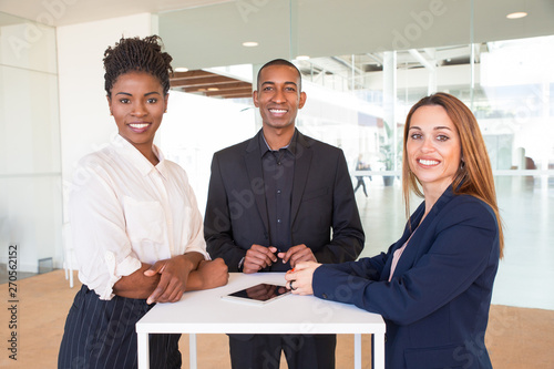 Cheerful successful team of three posing in office hallway. Business people in formal suits standing at counter and smiling at camera. Teamwork concept
