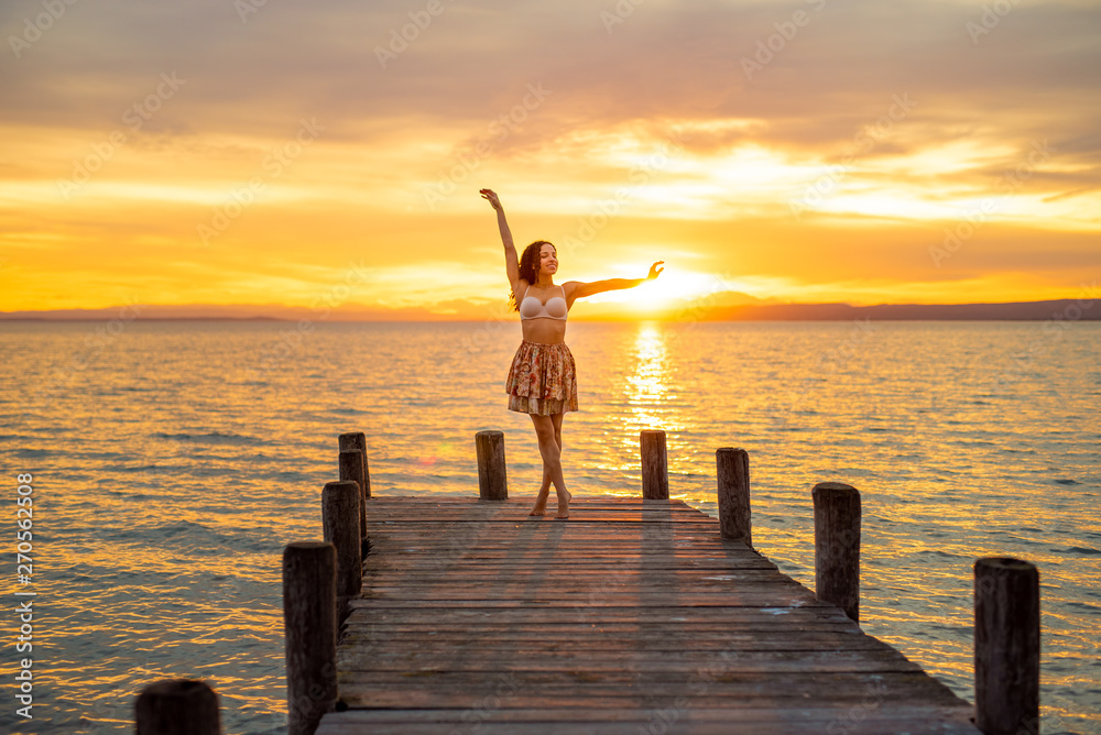 Fototapeta premium Happy woman enjoying the summery sunset at the lake on her vacation. The woman in bra and a short summer skirt laughs and puts her arms in the direction of the sky