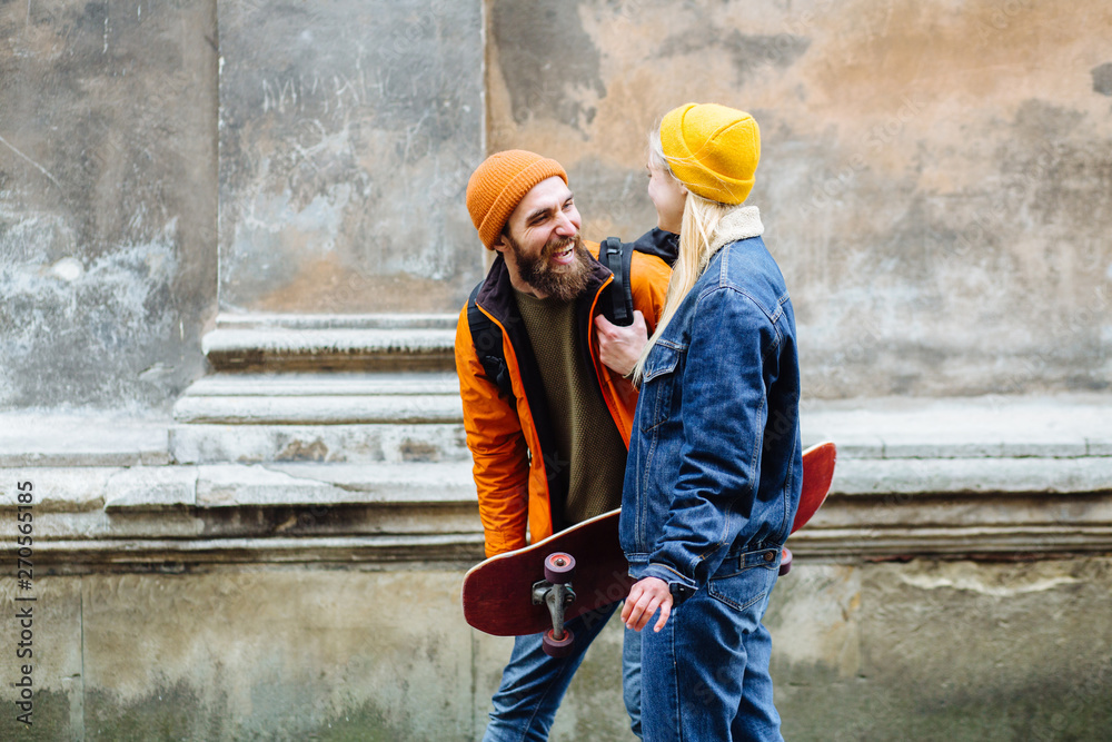 Happy young hipster couple traveler in bright casual clothes hold hands and walking together with skateboard over old wall on street in windy cold rainy weather outdoor. Happy young hipster couple traveler in bright casual clothes hold hands and walking together with skateboard over old wall on street in windy cold rainy weather outdoor.