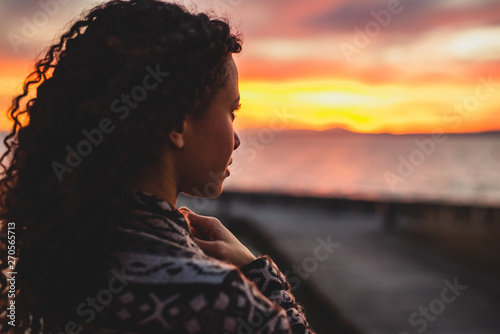 Thoughtful evening mood with a young Afro American woman standing on the promenade by the lake and looking towards the water and the setting sun. Burning sky