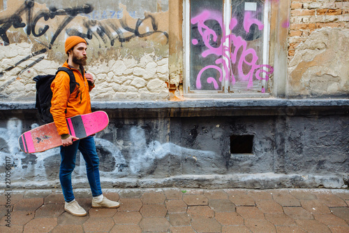 Hipster beard man traveler with longboard in his hand in orange hat and jacket black backpack walking in rainy urban old street. Full height.