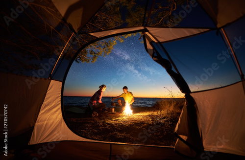 Camping on sea shore at sunset, view from inside tourist tent. Young couple hikers, man and woman preparing supper near campfire on blue sea water background. Tourism, recreation and love concept.