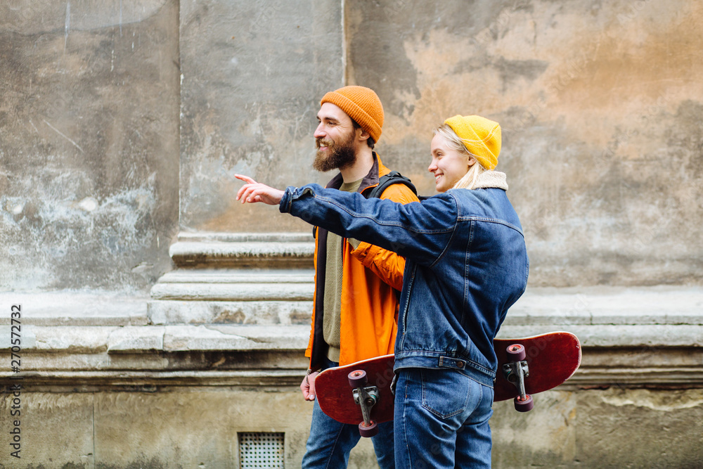 Happy young hipster couple traveler hold hands and walking together with skateboards over old wall on street in windy cold rainy weather outdoor.