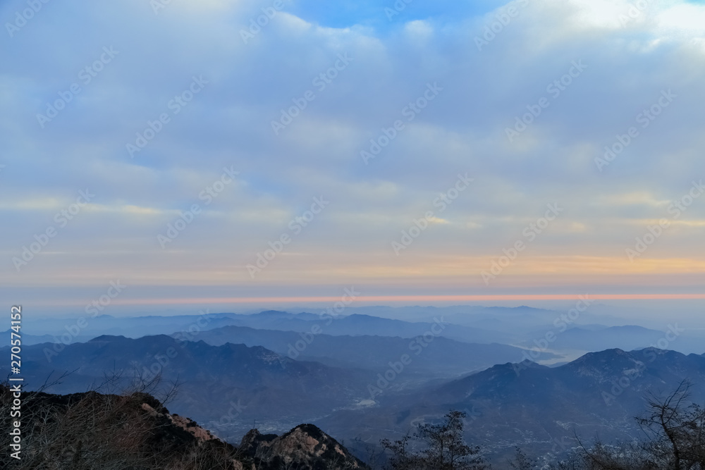 Rocks and snow on the peaks.Pine trees on the peak.The mountains in the distance loomed.Clouds wreathed it.The clouds in the sky were thick and heaving.Mountains piled up, clouds surging.