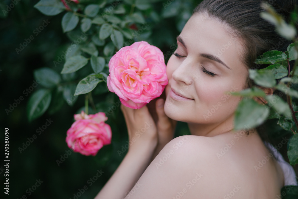 Young beautiful woman portrait close up with perfect skin posing with pink roses flowers in a garden