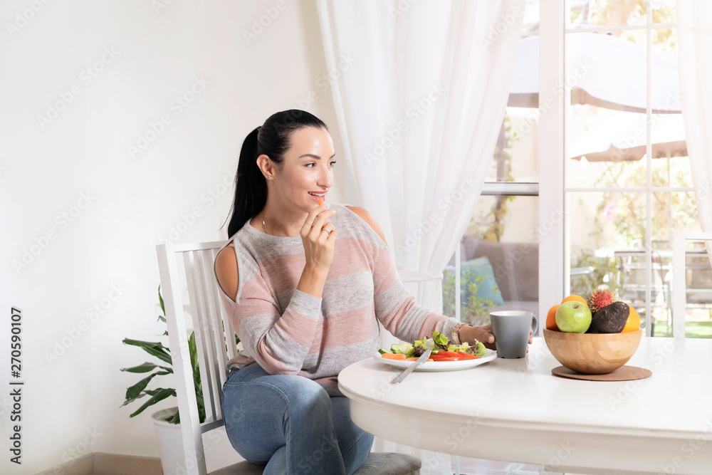 Beautiful brunette woman sitting at dining table eating a healthy salad and drinking coffee or tea with a bowl of fruits on table with garden in the background Beautiful brunette woman sitting at dining table eating a healthy salad and drinking coffee or tea with a bowl of fruits on table with garden in the background