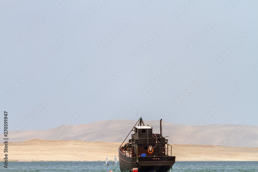 Fototapeta premium Old wooden boat sails the coast of Paracas (Peru) with dunes in the background