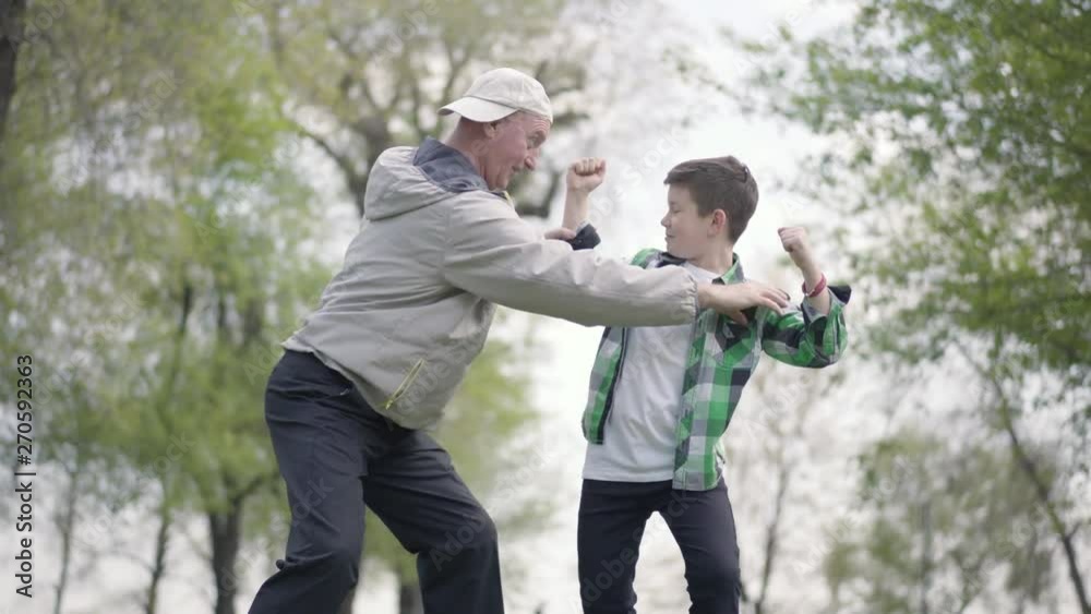The little boy in a checkered shirt and positive old man showing each other muscles in the park. Grandfather and grandson fooling around outdoors. Active lifestyle. Family leisure The little boy in a checkered shirt and positive old man showing each other muscles in the park. Grandfather and grandson fooling around outdoors. Active lifestyle. Family leisure