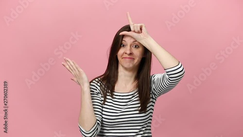 Smiling pretty young woman in striped shirt, fooling, showing loser gesture, pointing index finger on camera isolated over pastel pink background in studio. People sincere emotions lifestyle concept.