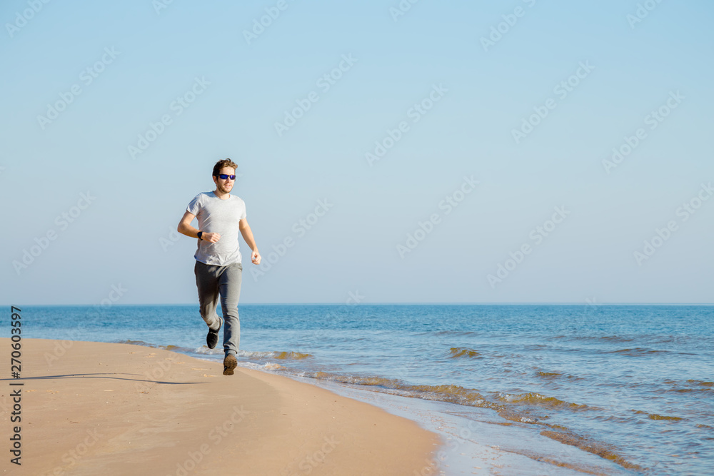 Young, adult alone man in t-shirt, sport trousers and sunglasses running on sand beach. Empty place for motivational, inspirational text, quote or sayings on light blue sky background. Front view. 