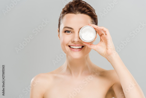 cheerful naked woman covering eye while holding container with face cream near eye and smiling isolated on grey