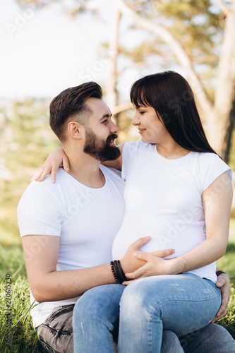 Happy young couple expecting baby, pregnant woman with husband touching belly, sitting on green grass