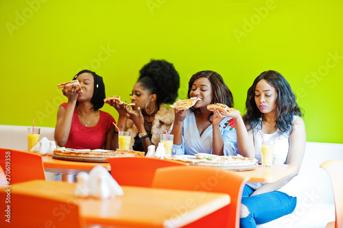 Four young african girls in bright colored restaurant eating pizza slices in hands.