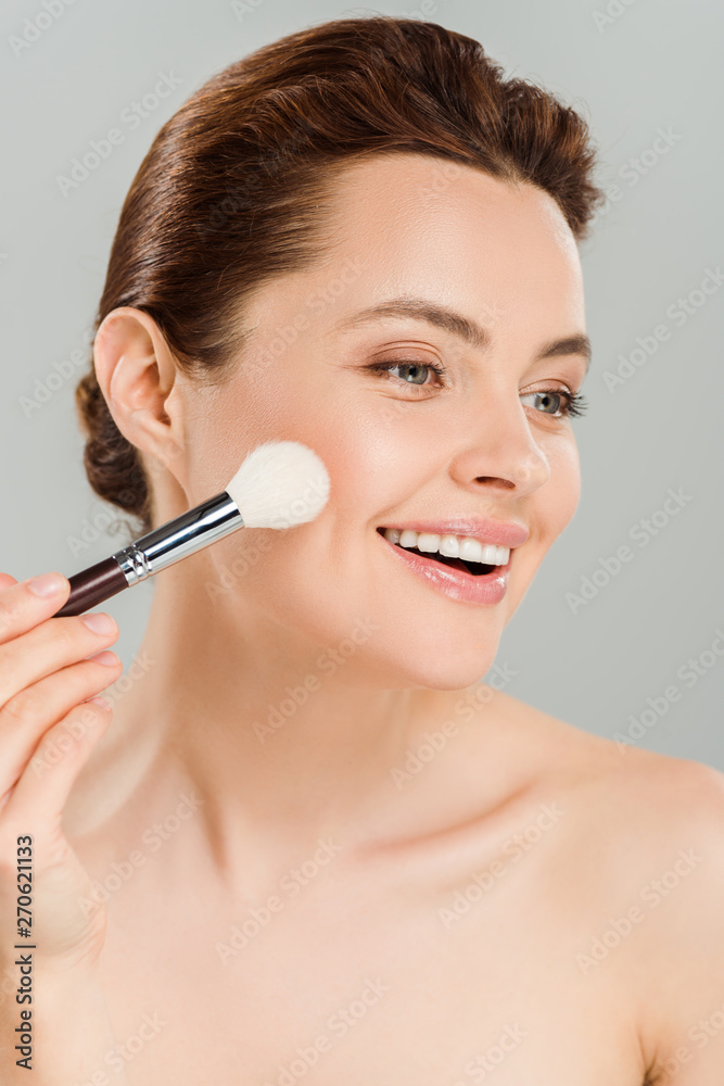 cheerful woman applying blush on cheek with cosmetic brush and smiling isolated on grey