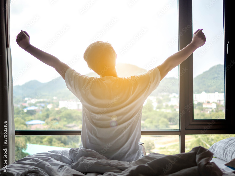 Man stretch his hands after waking up in bed at morning with sunlight - obrazy, fototapety, plakaty 