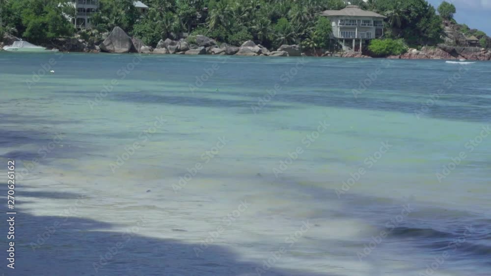 Seychelles. Praslin Island. Clear sea water rolls on the beach in waves with white foam close up. Small houses hidden in the tropical trees by the sea. Beautiful seascape. Camera moving up. Seychelles. Praslin Island. Clear sea water rolls on the beach in waves with white foam close up. Small houses hidden in the tropical trees by the sea. Beautiful seascape. Camera moving up.