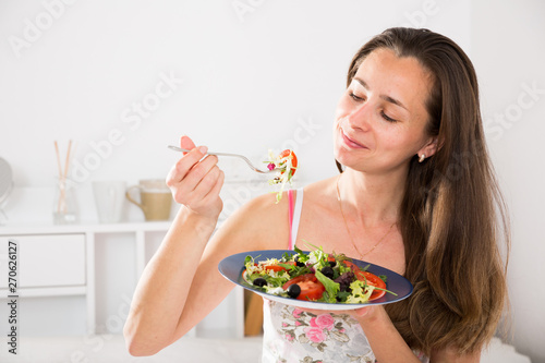 Positive pretty woman holding fork and eating vegetable salad in bed