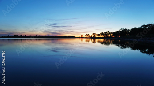 Sunset overlooking the lake, trees, clouds. The sky and bushes are reflected in the water. Landscape at dusk. Nightfall. Water like a mirror.