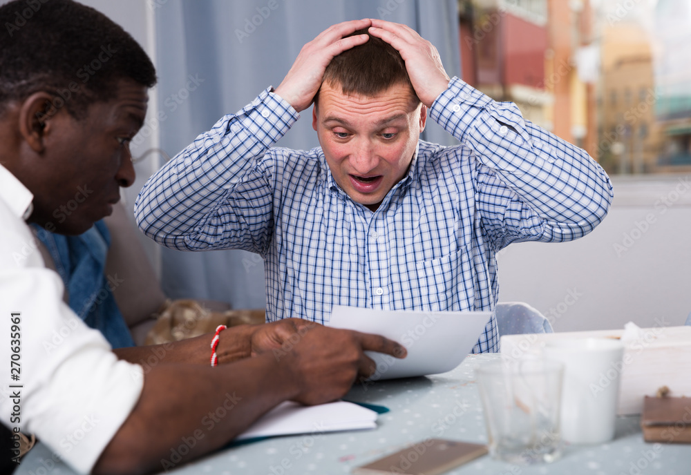 Upset man with friend looking worriedly at papers at home