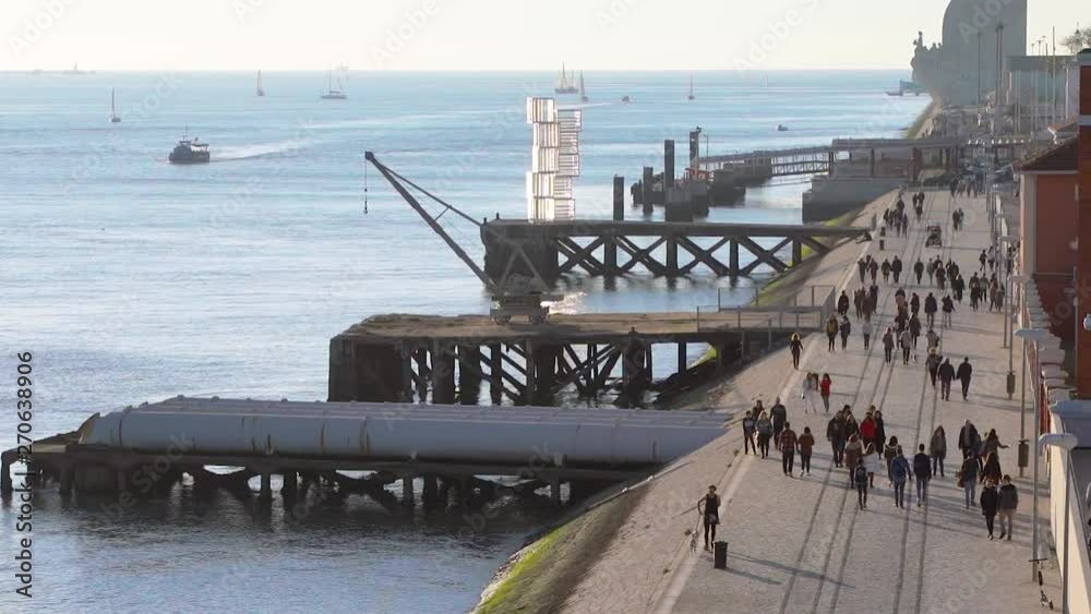 Pedestrian walkway riverside, tagus river. Monument of discovery, Padrao dos Descobrimentos, people passing by walking in slow motion with ocean view