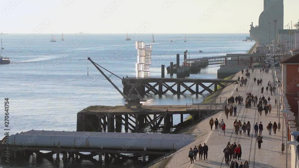 Pedestrian walkway riverside, tagus river. Monument of discovery, Padrao dos Descobrimentos, people passing by walking in slow motion with ocean view