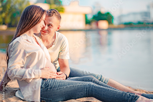 Beautiful pregnant woman and her husband are sitting on the bank of the river at sunset © ribalka yuli