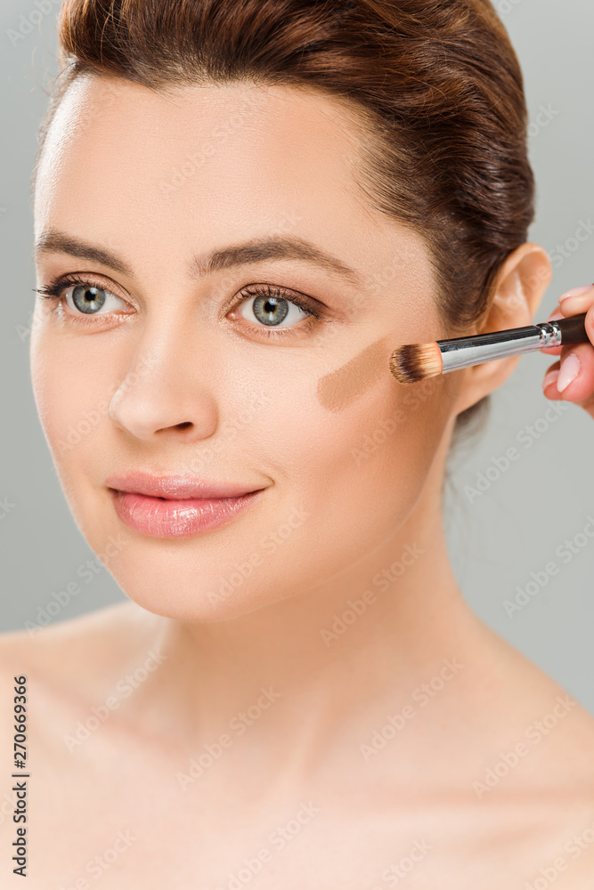 attractive woman holding cosmetic brush and applying brown blush on cheek isolated on grey