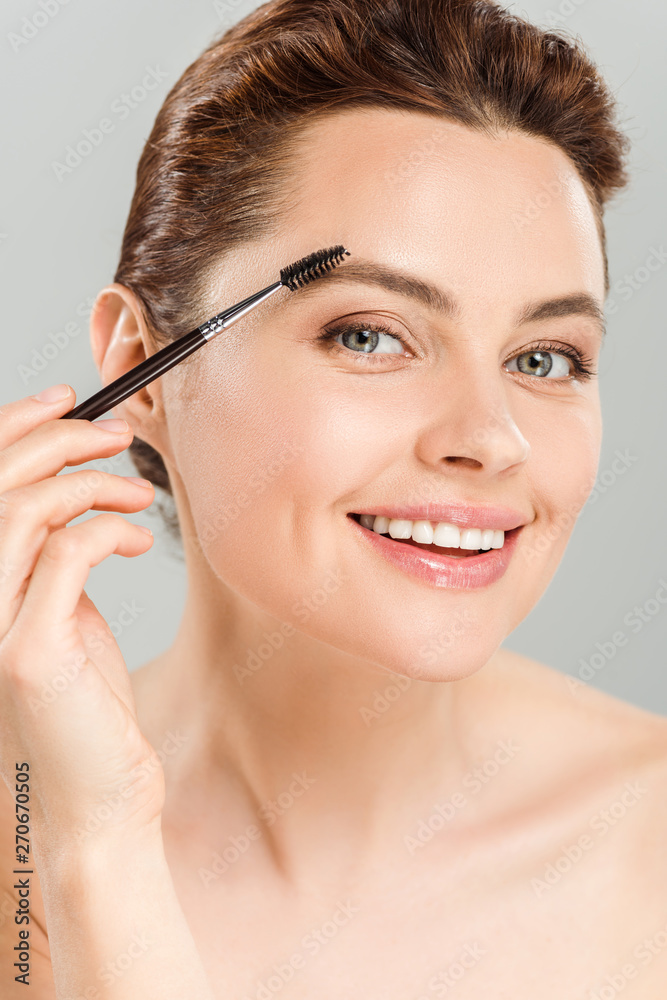 cheerful naked woman shaping eyebrow with eyebrow brush and smiling isolated on grey