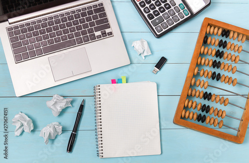 A calculator, a laptop and old abacus lie on a blue wooden table. The crumpled sheets speak of complex calculations. The link between generations.
